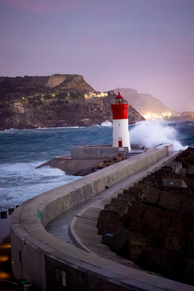 Lighthouse at the end of a concrete path with waves crashing against it during sunset.