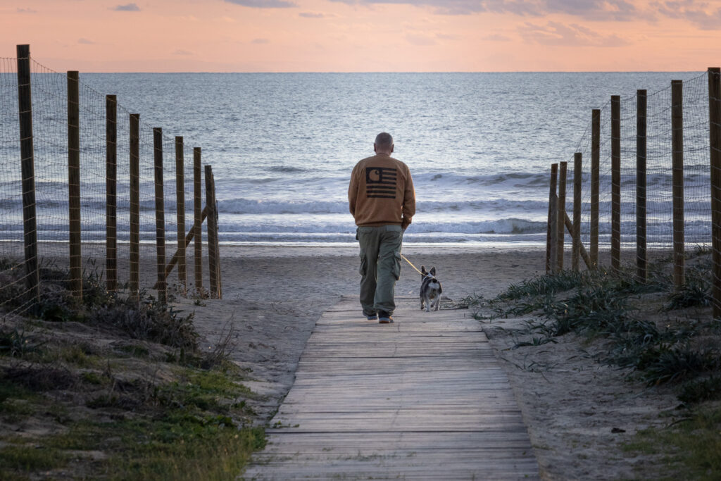 Man and dog walking on a wooden beach pathway at sunset, with symmetrical fences creating leading lines