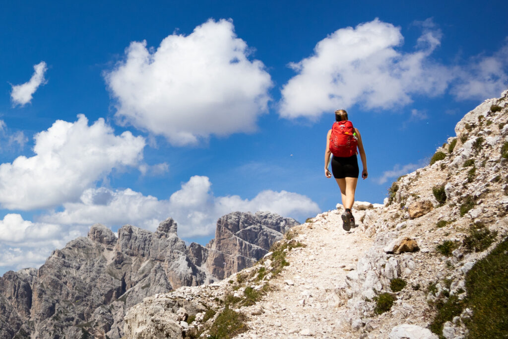 A woman with a red backpack hiking in the Alps, with a blue sky and mountains in the background.