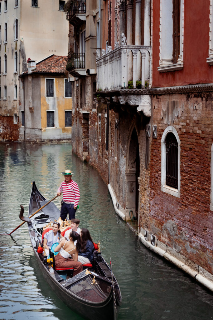 A man paddling a gondola with three passengers in Venice, with historic houses lining the riverbank.