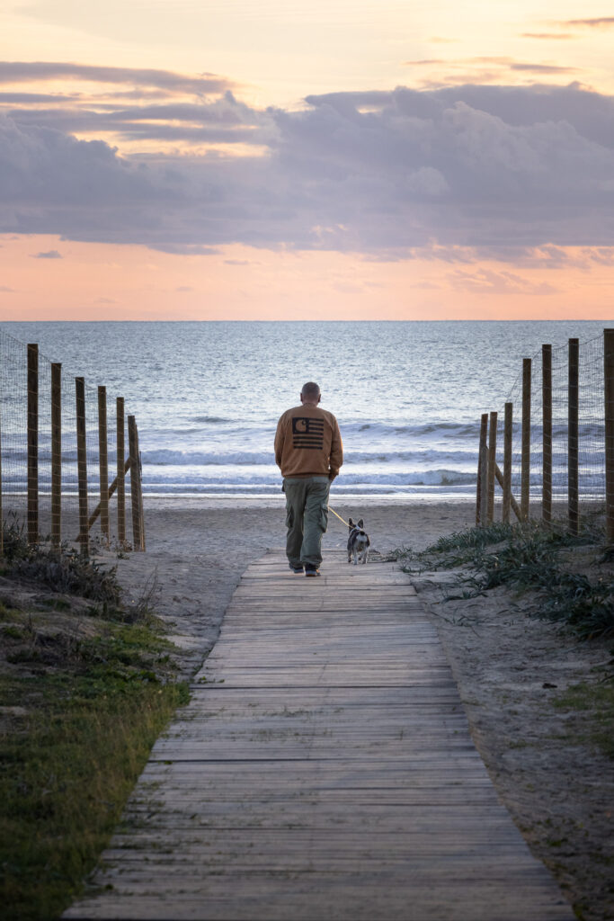 A man and a dog walking towards the sunset on a wooden pathway leading to the beach.